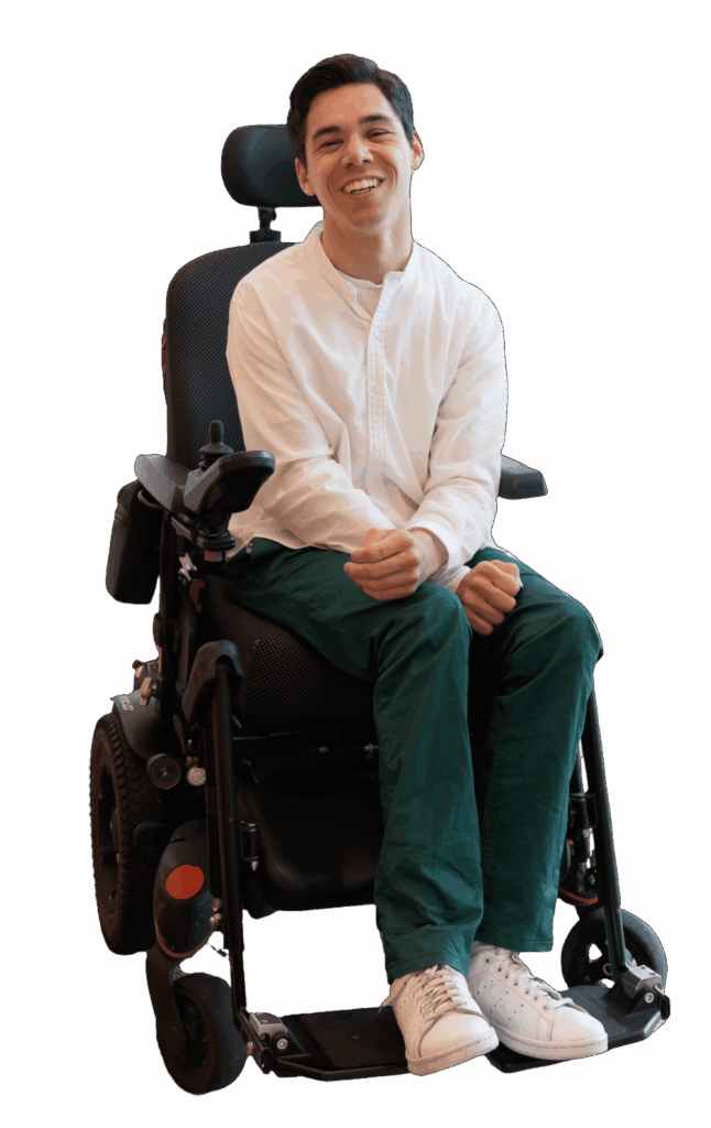 Maarten sitting in his electric wheelchair. He is wearing a white shirt, dark green pants, and white shoes. He is looking directly at the camera and smiling.